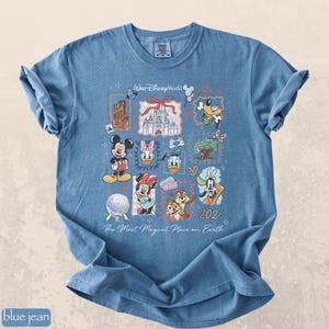 Op de afbeelding: Blauwe jeanskleurige t-shirt met een kleurrijke afbeelding van Disney-personages en bezienswaardigheden. Het ontwerp bevat Mickey Mouse, Minnie Mouse, Donald Duck en Goofy, samen met de tekst "Walt Disney World" en "2026". Het shirt heeft ook de tekst "The Most Magical Place on Earth".