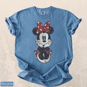 Pode incluir: Camiseta azul jeans com um gr&aacute;fico desgastado da Minnie Mouse. Minnie &eacute; retratada em preto e branco com um la&ccedil;o e detalhes vermelhos de bolinhas. A camiseta tem mangas enroladas e gola redonda.