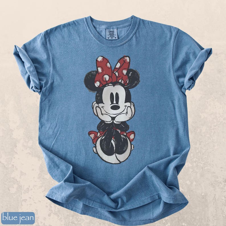 Pode incluir: Camiseta azul jeans com um gr&aacute;fico desgastado da Minnie Mouse. Minnie &eacute; retratada em preto e branco com um la&ccedil;o e detalhes vermelhos de bolinhas. A camiseta tem mangas enroladas e gola redonda.