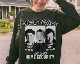 Puede incluir: Sudadera negra con un gráfico de tres personajes de la película "Solo en casa", el texto "McCallister's" y "Home Security Est. 1990". La sudadera tiene cuello redondo y mangas largas.