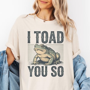 May include: Cream-coloured t-shirt with a detailed illustration of a toad and the text "I TOAD YOU SO" in dark grey. The toad is green and grey, with a textured appearance. The design is centered on the shirt.