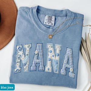 May include: A blue jean Comfort Colors t-shirt with the word "NANA" appliquéd across the front. The letters feature floral, polka dot, and gingham patterns. A gold necklace with a pendant is draped across the shirt.
