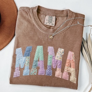 May include: A brown Comfort Colors t-shirt with the word "MAMA" appliquéd in patchwork fabric. The letters are in various pastel colours and patterns. A silver necklace with a pendant is draped on the shirt.