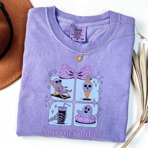 May include: A light purple Comfort Colors t-shirt with a graphic design featuring a skeleton enjoying summer activities. The design includes a bow, ice cream cone, drink, and ghost. The words "Summer Vibes" are printed at the bottom. A gold necklace is draped across the shirt.