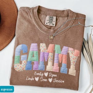 May include: Espresso-colored t-shirt with the word "GAMMY" appliqued in colorful patchwork letters. Below the name are the names "Emily", "Ryan", "Linda", "Sean", and "Jessica" with hearts. A silver necklace and a brown hat are visible.