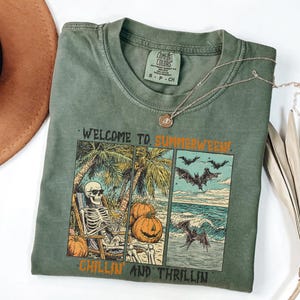 May include: Olive green t-shirt with a graphic that reads "WELCOME TO SUMMERWEED!" and "CHILLIN AND THRILLIN". The graphic features a skeleton in sunglasses relaxing on a beach, pumpkins, bats, and ocean waves. A silver necklace is draped across the shirt.