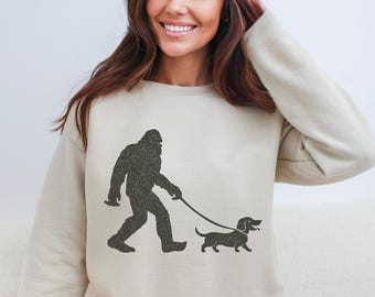 May include: Beige sweatshirt with a black graphic depicting Bigfoot walking a dachshund on a lead. The design has a distressed, vintage appearance. The sweatshirt is a neutral colour, suitable for casual wear.