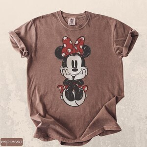 Pode incluir: Camiseta cor caf&eacute; com uma imagem da Minnie Mouse. Minnie &eacute; retratada em preto e branco com um la&ccedil;o e detalhes de bolinhas vermelhas. A camiseta tem gola redonda e mangas enroladas.