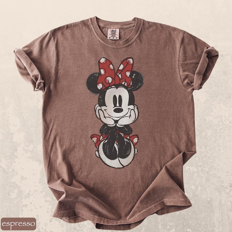 Pode incluir: Camiseta cor caf&eacute; com uma imagem da Minnie Mouse. Minnie &eacute; retratada em preto e branco com um la&ccedil;o e detalhes de bolinhas vermelhas. A camiseta tem gola redonda e mangas enroladas.