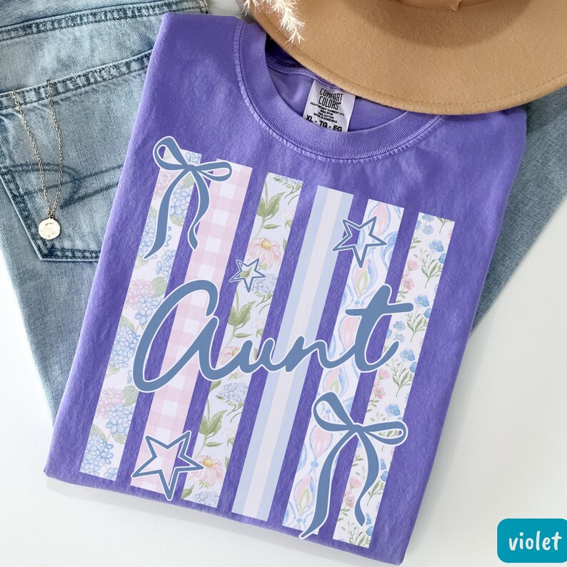 May include: A violet Comfort Colors t-shirt with the word "Aunt" in a cursive font. The shirt features vertical stripes with floral, gingham, and star designs. A light brown hat and blue jeans are in the background.