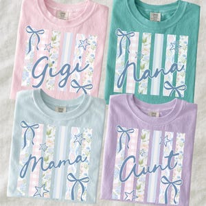 May include: Four pastel-colored t-shirts with the names "Gigi", "Nana", "Mama", and "Aunt" in cursive script. Each shirt features a vertical design with floral, gingham, and striped patterns, accented with bows and stars. The shirts are light pink, teal, light blue, and lavender.