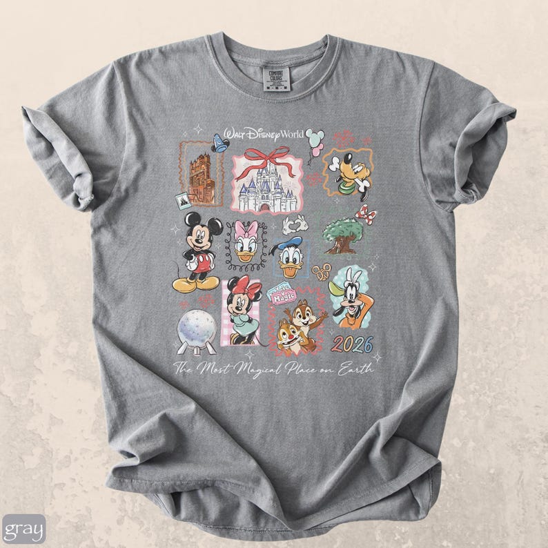 Op de afbeelding: Grijs T-shirt met een kleurrijk Disney-thema ontwerp. Het ontwerp bevat Mickey Mouse, Minnie Mouse, Donald Duck en Goofy, samen met de tekst "Walt Disney World" en het jaar "2026". De zin "The Most Magical Place on Earth" is ook aanwezig.