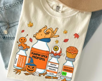 May include: Cream-colored Comfort Colors t-shirt with a Halloween-themed graphic. The design features cartoon vials labeled "Pumpkin Spice Propofol," "Maple Cinnamon Haldol," and "Spiced Vanilla Versed." Includes a witch's hat, a pumpkin, and fall leaves.