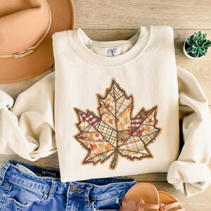 May include: A cream-coloured sweatshirt with a large, patchwork-style leaf design in autumnal colours. The leaf design incorporates various patterns, including plaid and floral prints. The sweatshirt is paired with blue denim shorts and a brown hat.