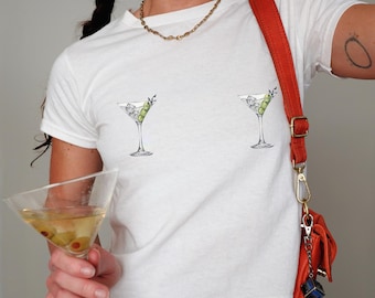 May include: White t-shirt featuring two martini glass illustrations with olives, ice, and a sprig of greenery. The shirt has a classic crew neck and short sleeves. A gold chain necklace and a red shoulder bag with a blue charm are also visible.