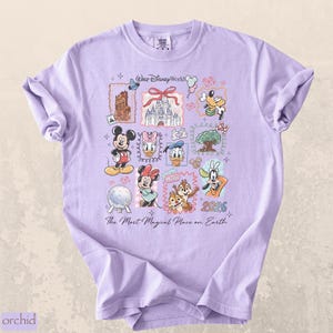 Op de afbeelding: Een lichtpaars t-shirt met een kleurrijk grafisch ontwerp met Disney-personages en bezienswaardigheden. Het ontwerp bevat Mickey Mouse, Minnie Mouse, Donald Duck en Goofy, samen met de tekst "Walt Disney World" en "2026".