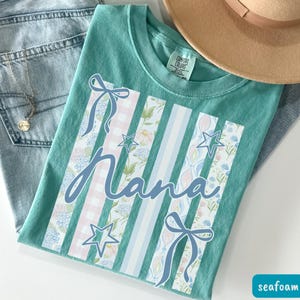 May include: Seafoam green t-shirt with the word "Nana" in a cursive font. The shirt features vertical stripes with floral, gingham, and star patterns. A light brown hat and blue jeans are in the background.