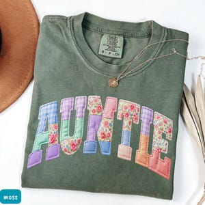 May include: Olive green Comfort Colors t-shirt with the word "AUNTIE" appliqued across the front in colorful patchwork fabric. The letters feature floral, plaid, and solid patterns. A delicate gold necklace with a pendant rests on the shirt.
