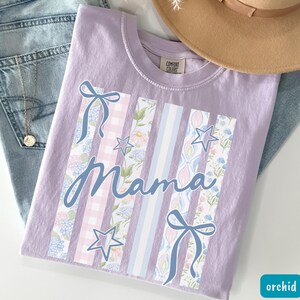 May include: Orchid-colored t-shirt with the word "Mama" in cursive script. The shirt features vertical stripes with floral, gingham, and striped patterns, along with blue bows and stars. The shirt is folded on top of a pair of jeans.