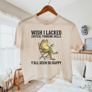 Könnte beinhalten: Ein cremefarbenes Kurzarm-T-Shirt mit einer Grafik eines grünen Frosches, der in einem Klappstuhl sitzt und eine Tasse hält. Der Text über dem Frosch lautet WISH I LACKED CRITICAL THINKING SKILLS und der Text darunter lautet Y'ALL SEEM SO HAPPY.