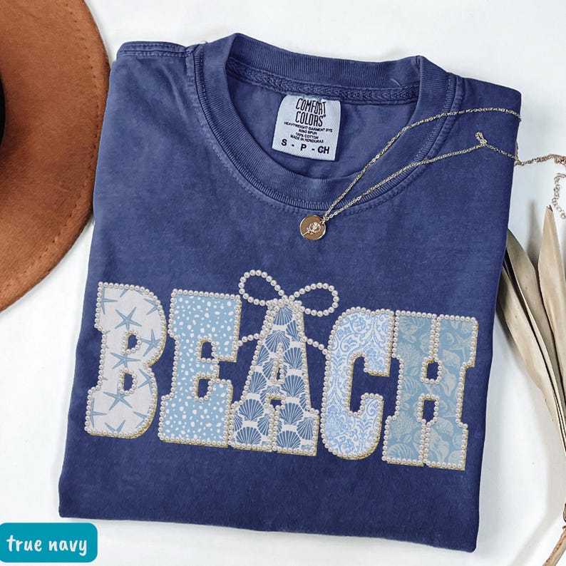 May include: A true navy blue t-shirt with the word "BEACH" in a decorative design. The letters are light blue and white, with seashell and starfish patterns. A gold necklace with a pendant is draped across the shirt.