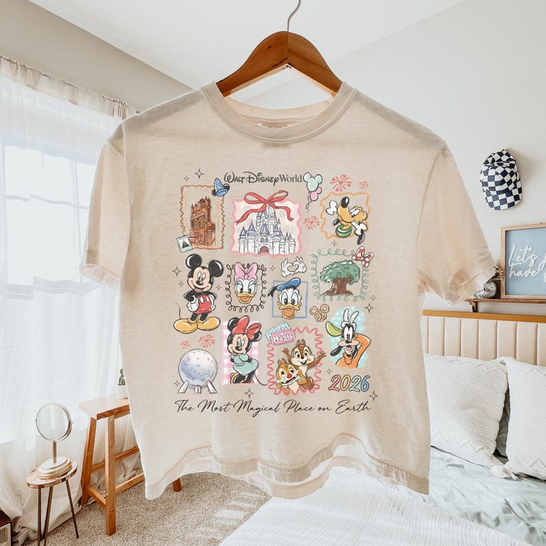 Op de afbeelding: Beige T-shirt met korte mouwen met een kleurrijke afbeelding van Disney-personages en bezienswaardigheden. Het ontwerp bevat Mickey Mouse, Minnie Mouse, Donald Duck en Goofy, samen met de tekst "Walt Disney World" en "2026". Het shirt heeft ook de tekst "The Most Magical Place on Earth".