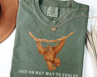 Op de afbeelding: Saliegroen t-shirt met een orang-oetan afbeelding die aan een tak hangt. De tekst "JUST ON MAY WAY TO EVOLVE" staat onderaan gedrukt. Een zilveren ketting met een hanger ligt over het t-shirt.