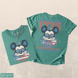 Puede incluir: Dos camisetas verde claro con un gr&aacute;fico de Mickey Mouse con gafas de sol con un dise&ntilde;o patri&oacute;tico. Las camisetas tienen el texto "1776" y "United States Independence Day". Una camiseta tiene un collar de cadena plateada.