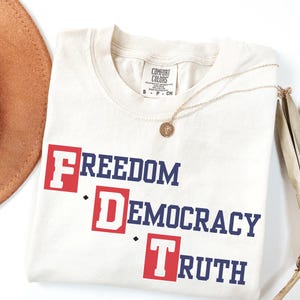 May include: Cream-colored t-shirt with the words "FREEDOM DEMOCRACY TRUTH" in a bold, patriotic design. The text is in a mix of red and blue. A gold necklace with a pendant rests on the shirt. The shirt has a Comfort Colors tag.