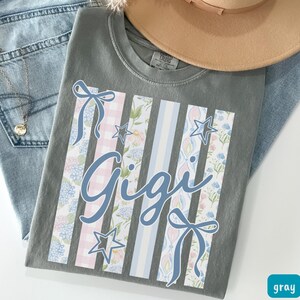 May include: Gray t-shirt with the name "Gigi" in cursive script. The shirt features vertical stripes with floral, gingham, and star patterns in pink, blue, and white. A light brown hat and blue jeans are in the background.