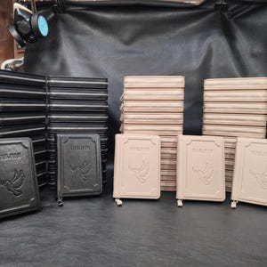 May include: A collection of leather-bound books with the word "БІБЛІЯ" embossed on the cover, featuring a dove design. The books are in various colors, including black and beige, with zippered closures. Stacks of similar books are visible.