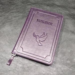 Puede incluir: Una Biblia morada con la palabra "БИБЛИЯ" en relieve en la portada. Una paloma con una rama de olivo también está en relieve en la portada. La Biblia tiene cierre de cremallera y es de material similar al cuero.