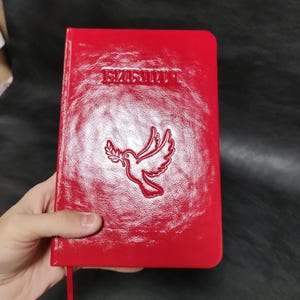 Puede incluir: Un libro rojo con texto en relieve y una paloma que lleva una rama de olivo. La portada del libro tiene la palabra "БИБЛИЯ" en relieve en la parte superior. Una cinta roja de marcador es visible.