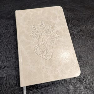 Puede incluir: Un diario beige con un diseño en relieve de un corazón humano con elementos florales. La cubierta tiene una apariencia texturizada similar al cuero. Una cinta blanca como marcapáginas es visible. El diario es rectangular con esquinas redondeadas.