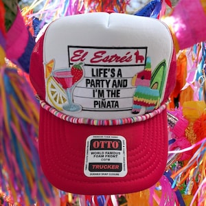 May include: A red and white trucker hat with the text "El Estrés, Life's a Party and I'm the Piñata." The hat features a cocktail and piñata graphic. It has a foam front and a snap closure.