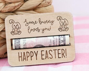 Op de afbeelding: Houten paas cadeaubonnenhouder met gegraveerde konijnen illustraties en de tekst "Some bunny loves you!" en "HAPPY EASTER!". Een opgerolde bankbiljet is zichtbaar in de gleuf. De achtergrond heeft een gevlochten mand en een roze en wit geblokt patroon.