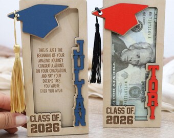 Op de afbeelding: Twee houten afstudeergeschenkhouders. Eén heeft een blauwe baret, de naam "Julian" in blauw en de tekst "Klasse van 2026". De andere heeft een rode baret, de naam "Tori" in rood en de tekst "Klasse van 2026". Beide hebben een kwast.