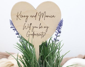 Op de afbeelding: Een houten hartvormig bord met de tekst "Kenny and Monica, Will you be my Godparents?" wordt weergegeven in een pot met groen. Het bord is lichtbruin met zwarte letters. Paarse bloemen zijn zichtbaar.