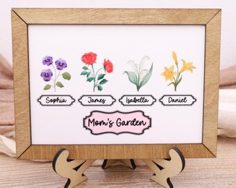 Op de afbeelding: Een ingelijste print met een houten frame en standaard toont aquarelbloemen en namen: Sophia, James, Isabella en Daniel. De tekst "Mom's Garden" staat onder de namen. Het frame wordt ondersteund door een houten standaard.