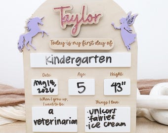 Op de afbeelding: Een boogvormig houten bord met de naam Taylor, een paarse eenhoorn en een fee. Het bevat de tekst Today is my first day of Kindergarten, met witte vakken voor Date: Aug 18, 2026, Age: 5, Height: 43", a veterinarian, en unicorns, fairies, ice cream.