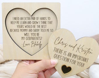 Op de afbeelding: Twee houten hartvormige plaquettes. Eén heeft een hartvormige uitsparing en de tekst: "Ik heb een extra paar handen nodig... Willen jullie mijn peetouders zijn? Love Blakely." De andere zegt: "Chris en Kristen, er is een belangrijke vraag voor jullie!"