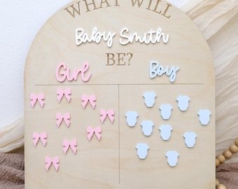 May include: A wooden gender reveal sign with the text "WHAT WILL Baby Smith BE?" in a light brown color. The sign is divided into two sections, one for "Girl" with pink bows and one for "Boy" with blue onesies.
