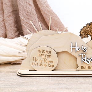 May include: Wooden Easter decoration with a sun, a tree, and a stone tomb. The tomb is open and the text "He is not here for He is Risen just as He said" is engraved on a round plaque. The text "He is Risen" is engraved on a separate plaque with the scripture reference "Matthew 28:6".