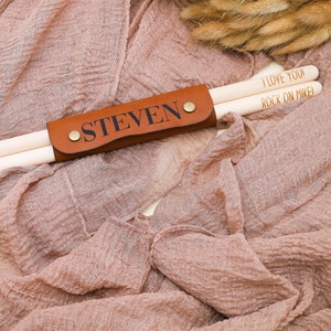 May include: A brown leather drumstick holder with the name "STEVEN" engraved on it. The holder is holding two wooden drumsticks with the text "I LOVE YOU" and "ROCK ON MIKE!" written on them.