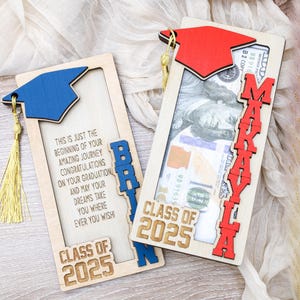 May include: Two wooden graduation card holders with a slot for cash. The card holders are decorated with a graduation cap and the text "Class of 2025". One card holder is blue and says "Brian" and the other is red and says "Makayla".