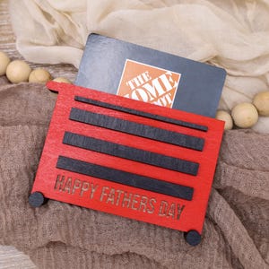 May include: A red wooden gift card holder shaped like a toolbox, with black accents and the words "HAPPY FATHERS DAY." A gift card is inserted into the holder. The background includes a neutral-toned fabric and wooden beads.