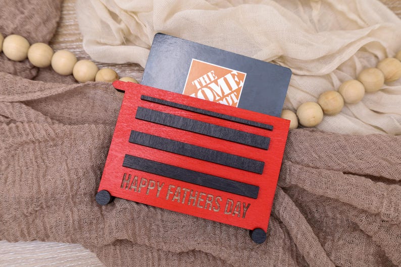May include: A red wooden gift card holder shaped like a toolbox, with black accents and the words "HAPPY FATHERS DAY." A gift card is inserted into the holder. The background includes a neutral-toned fabric and wooden beads.