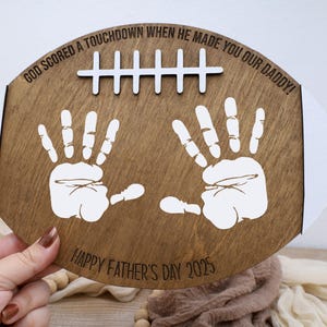 May include: A wooden football-shaped plaque with white handprints and the text "GOD SCORED A TOUCHDOWN WHEN HE MADE YOU OUR DADDY!" and "HAPPY FATHER'S DAY 2025". The plaque has white stripes and a white design resembling football laces.