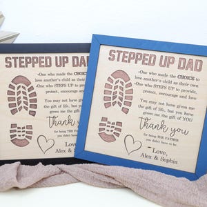 May include: Two wooden signs with blue and black frames. The signs have a wood grain background with a brown footprint design and the text "Stepped Up Dad". The text reads: "One who made the choice to love another's child as their own. One who steps up to provide, protect, encourage and love. You may not have given me the gift of life, but you have given me the gift of you. Thank you for being the father you didn't have to be. Love, Alex & Sophia."