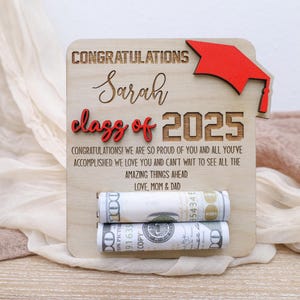 May include: Wooden sign with a red graduation cap, the text "Congratulations Sarah Class of 2025" and a message of congratulations. The sign has two rolled up dollar bills tucked into slots.